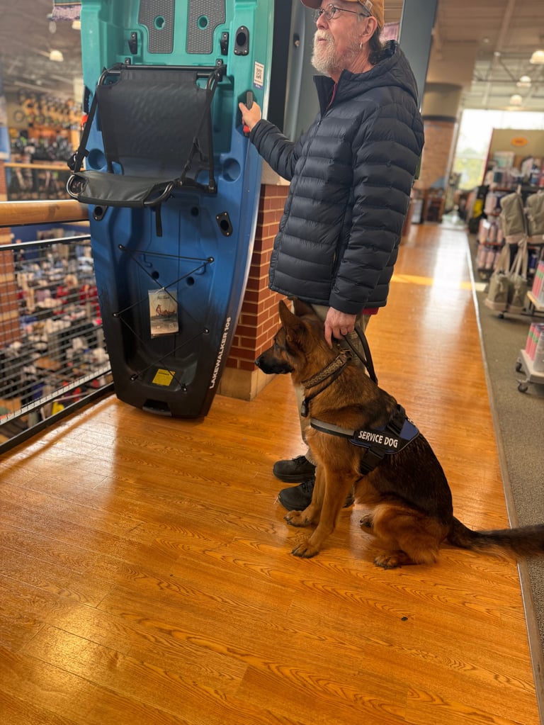 Man in black puffer jacket standing next to a blue robotic device with a brown dog sitting on wooden floor in a retail space