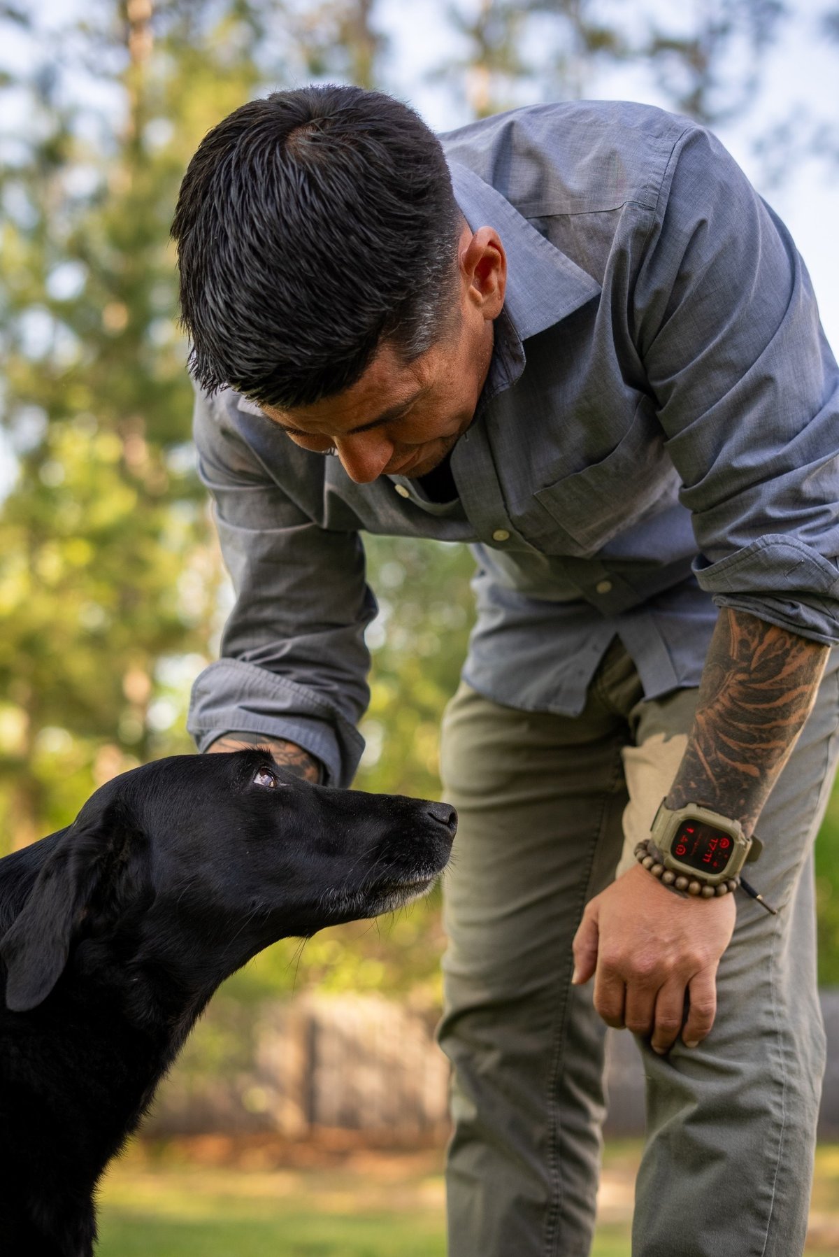 Man bending down to pet a black dog in an outdoor garden setting with blurred foliage in background