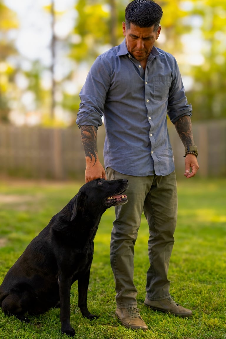 Man in blue shirt standing with black dog on grass in backyard with blurred green foliage